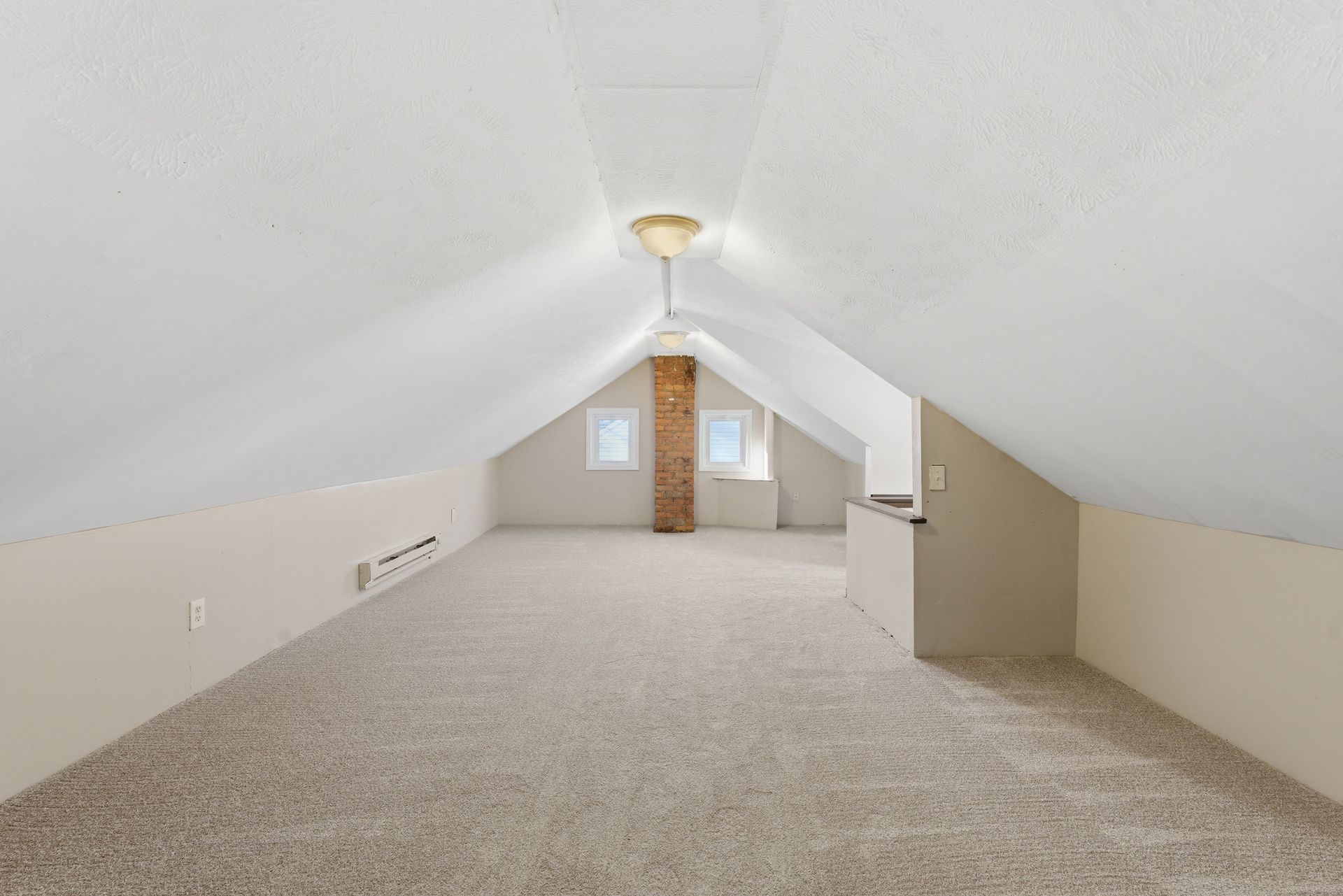 Empty attic room with sloped white ceilings, carpeted floor, and a central brick chimney.