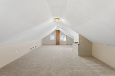 Empty carpeted attic room with sloped white ceiling, skylights, and central brick chimney.