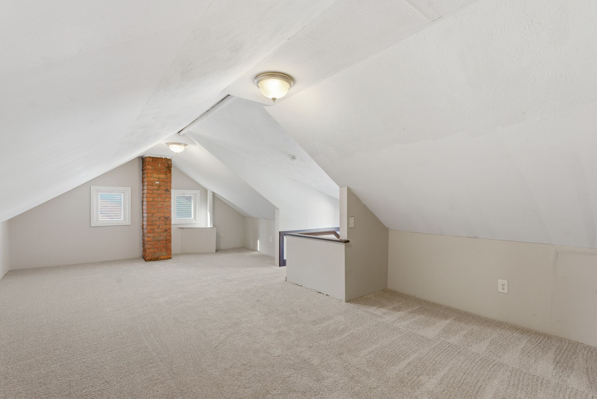 Empty carpeted attic room with white sloped ceilings, a brick chimney, and a small window.