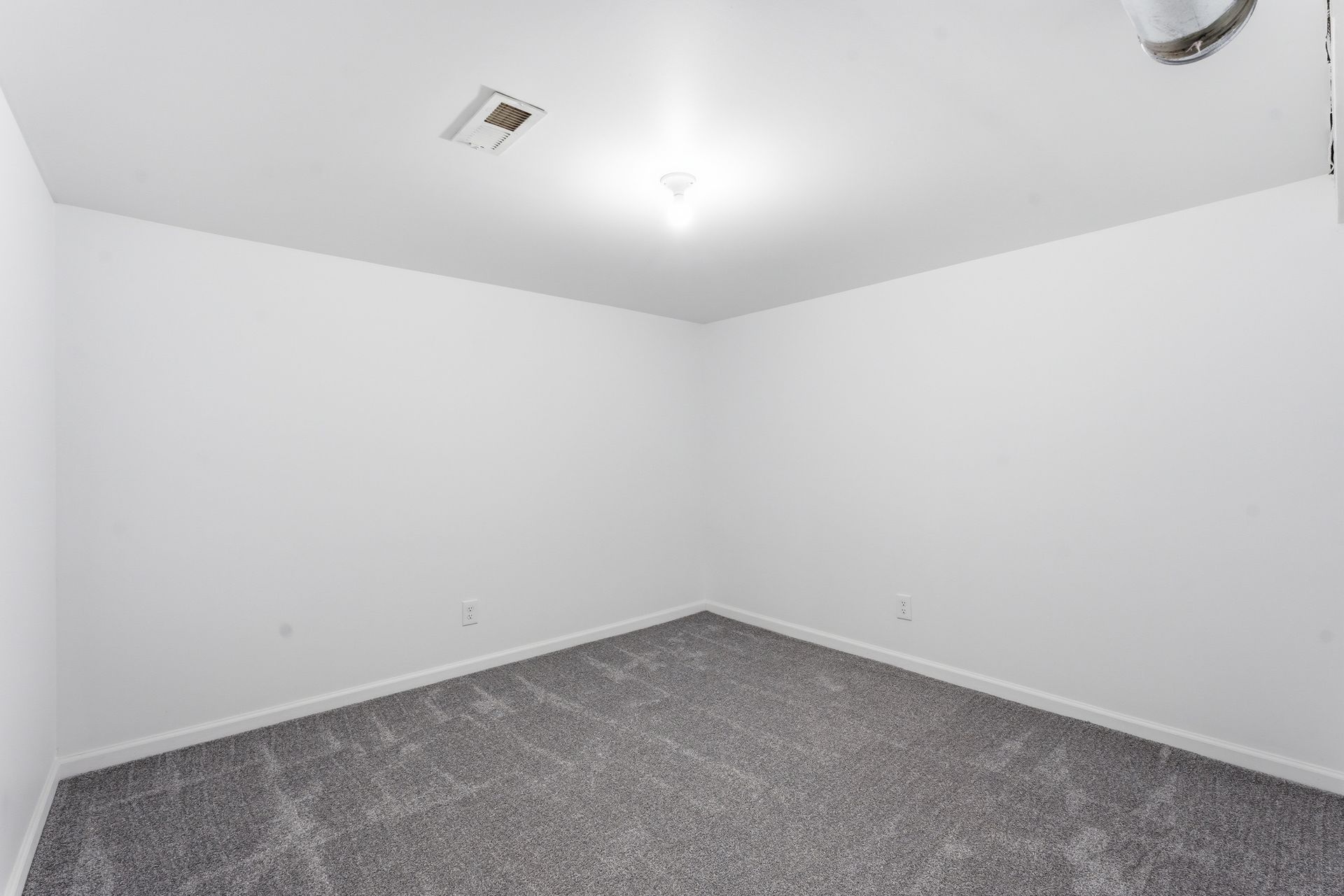 Empty white room with gray carpet and ceiling light, viewed from a corner.