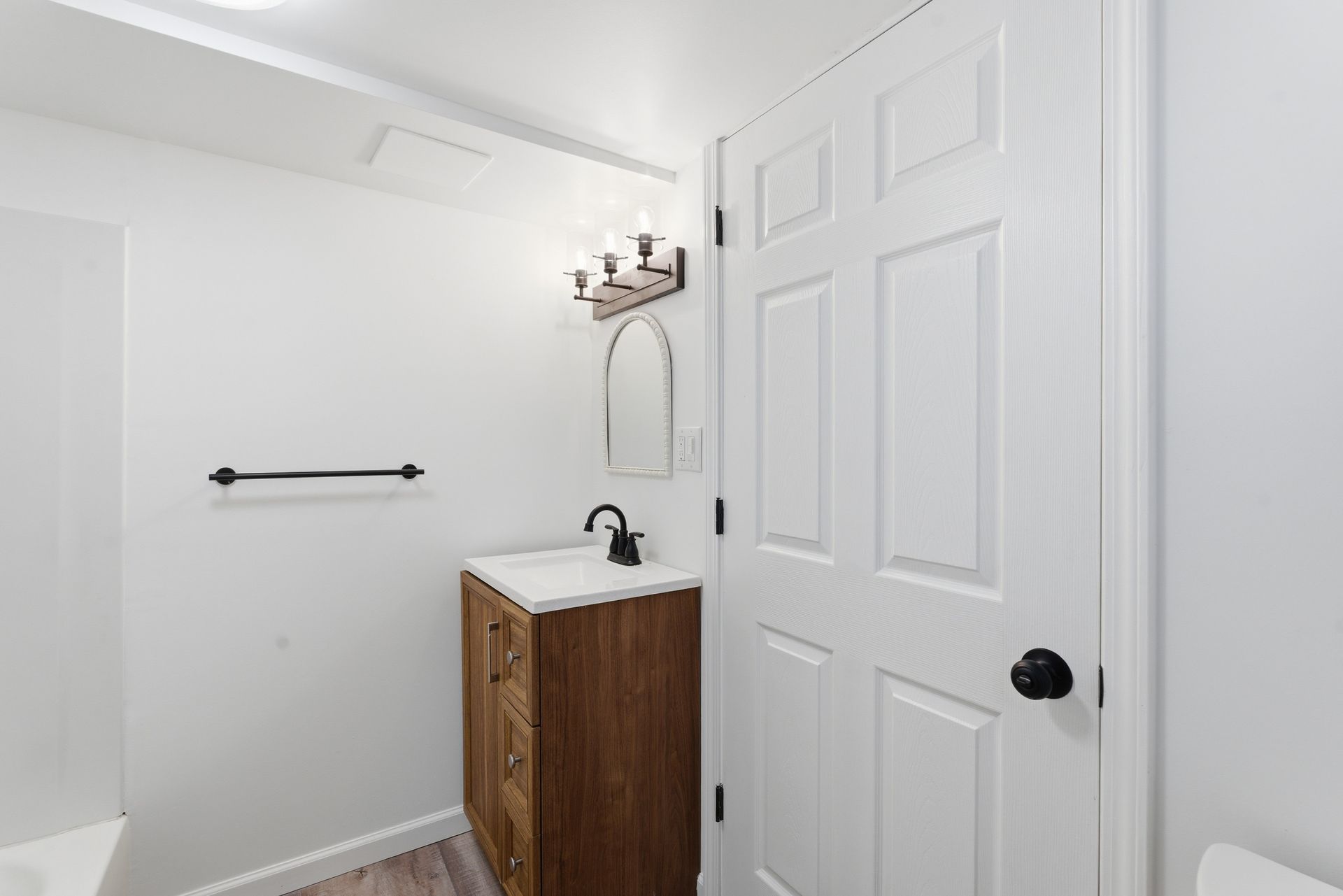 Small white bathroom with a wood vanity, wall mirror, towel bar, and an open door.