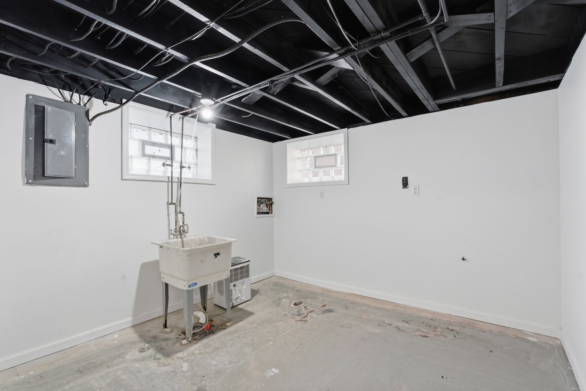 Empty basement utility room with white walls, exposed black ceiling joists, small windows, and a sink basin.