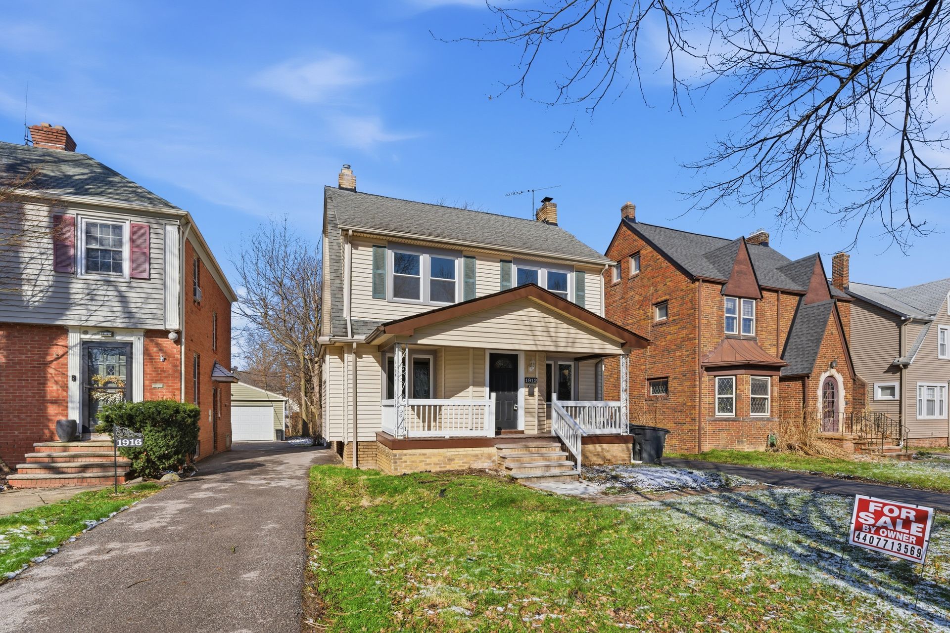 Detached two-story brick house with a front porch, driveway, and lawn on a sunny day