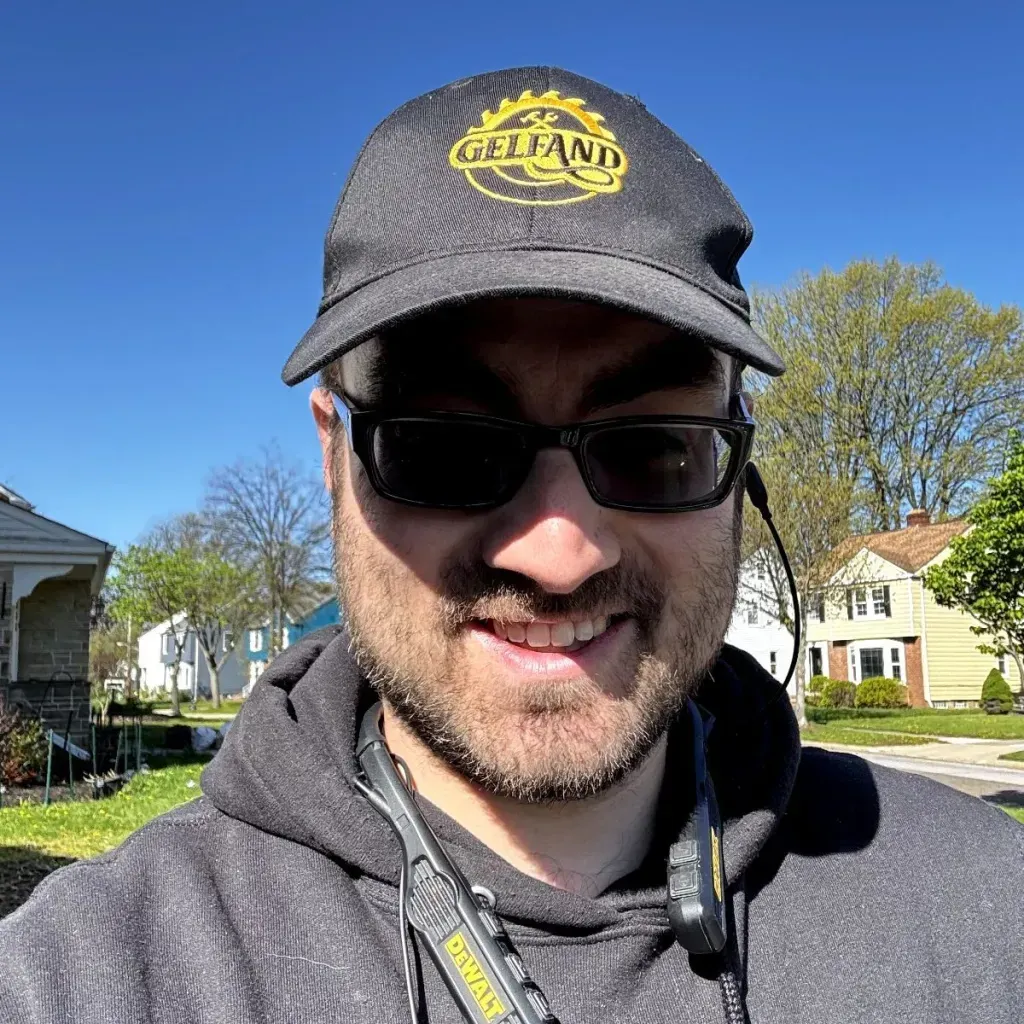 Man in sunglasses and hat smiles outdoors on a sunny day. Houses and trees are in the background.