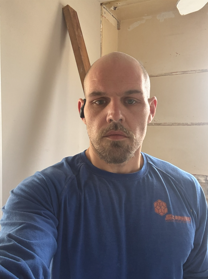 Person in a blue shirt taking a selfie in an unfinished room with exposed walls.