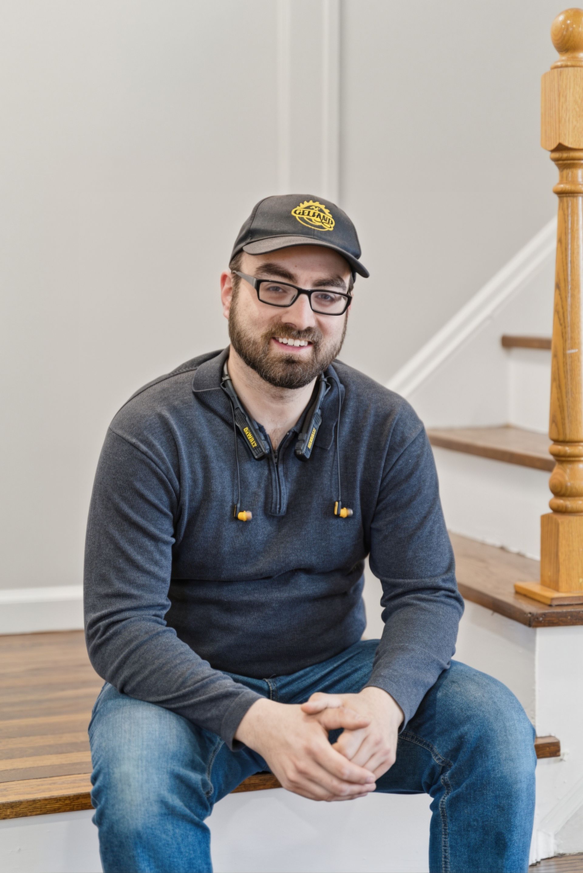Man seated on indoor stairs, wearing a cap and glasses, smiling at the camera