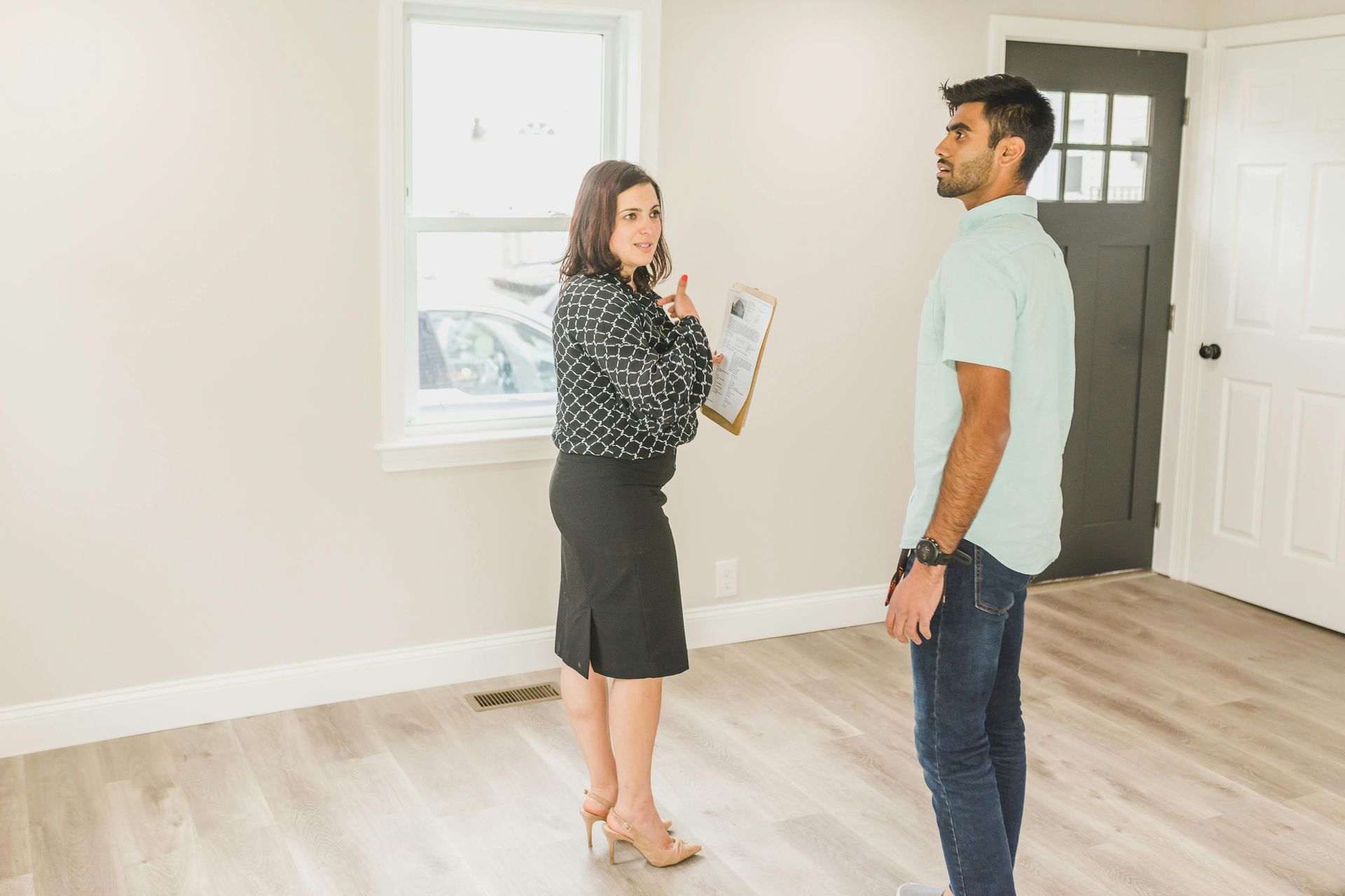 Two people talking in a bright empty room, one holding a clipboard and pointing toward the window.