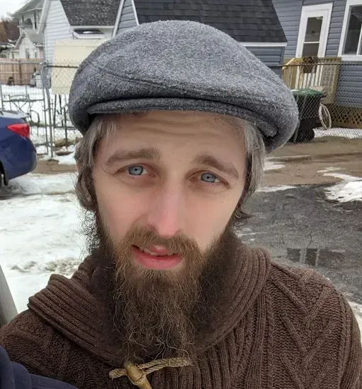 Man with blue eyes and graying hair, wearing a gray cap and brown sweater; snowy outdoor setting.