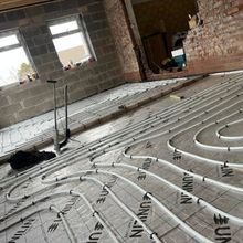 Underfloor heating installation in a room with exposed brick and concrete block walls. White pipes are laid on insulated underlay.