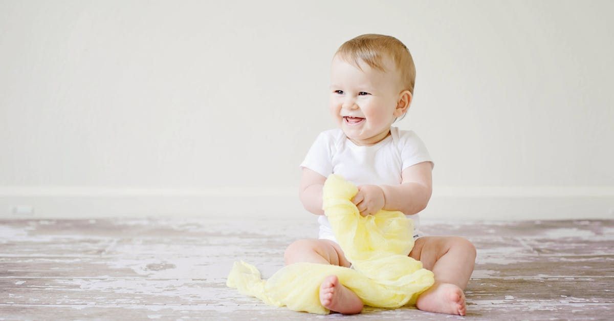 A smiling baby sitting on a carpeted floor holding a small yellow blanket. 