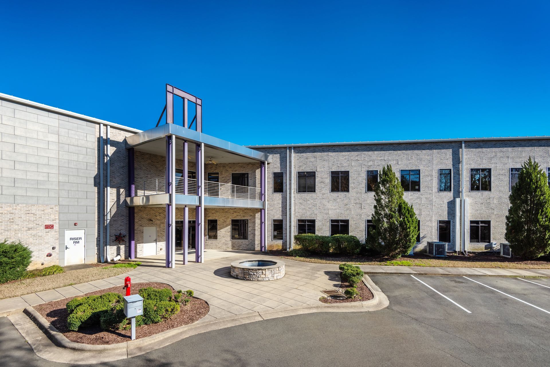 A large brick building with a purple balcony is surrounded by trees and a parking lot.