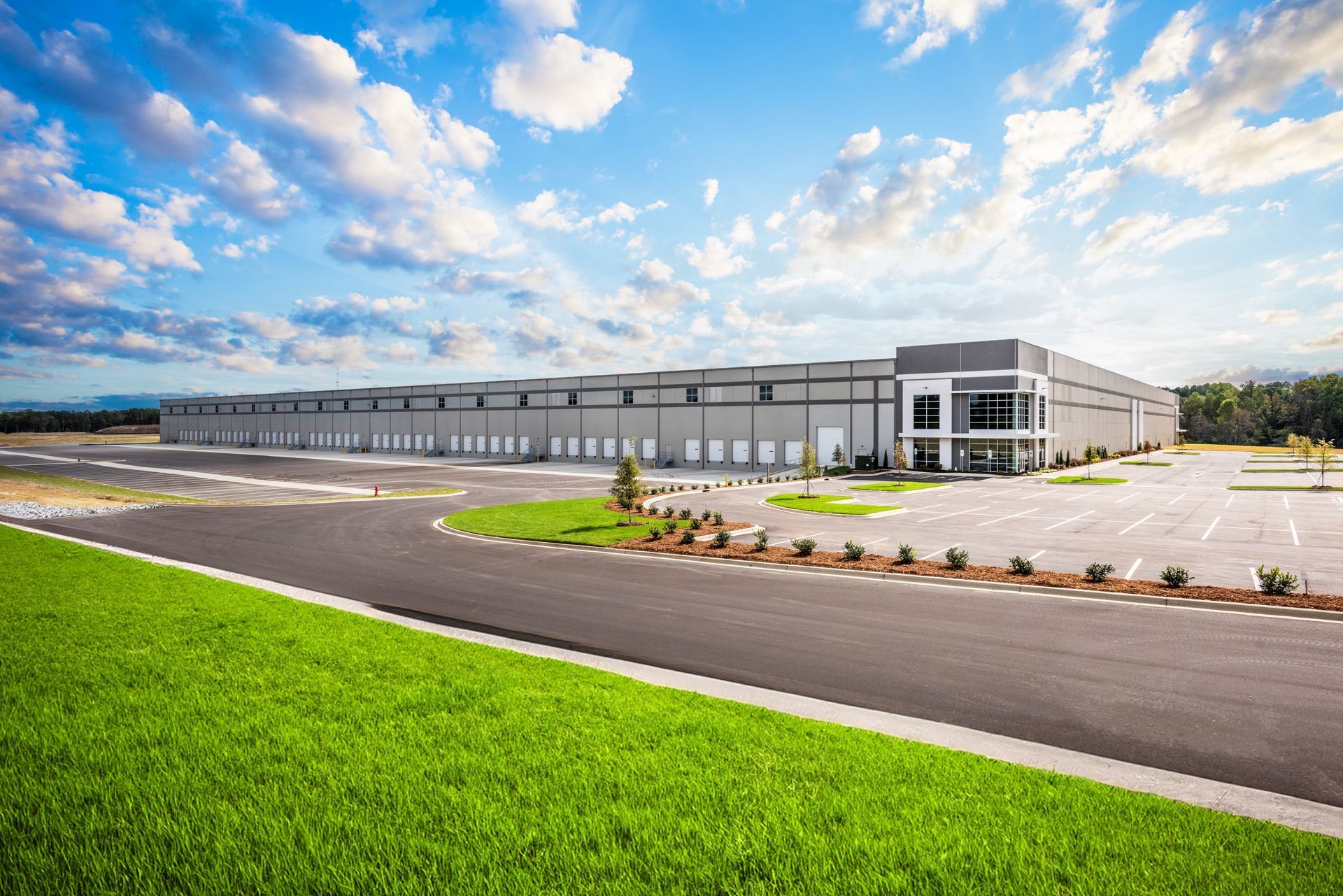 A large warehouse is sitting on top of a lush green field next to a road.