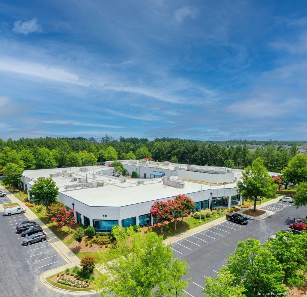 Aerial view of a white commercial building with blue accents, surrounded by parking and lush green trees under a bright blue sky.