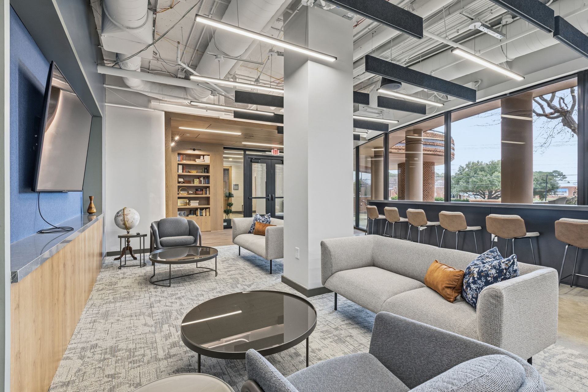 Modern office lounge area with gray sofas, a round coffee table, a tall pillar, and bar stools facing a large window.