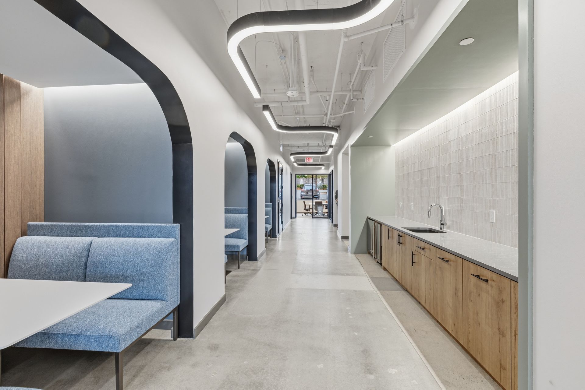 A hallway in a modern office with blue bench seating in arched alcoves on the left and a wood-cabinet kitchenette on right.