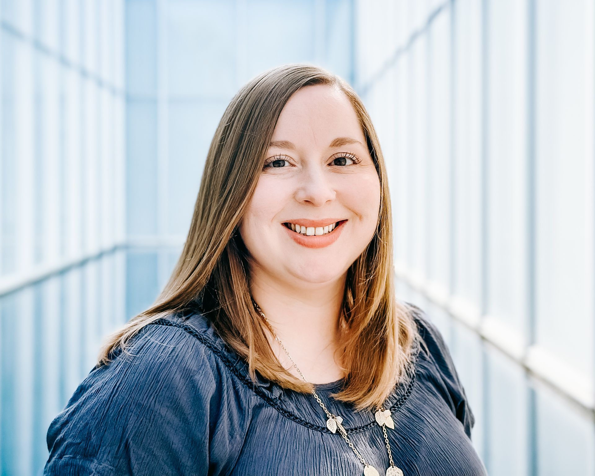 A smiling woman with long brown hair wearing a dark blue blouse and a delicate necklace against a light blue backdrop.