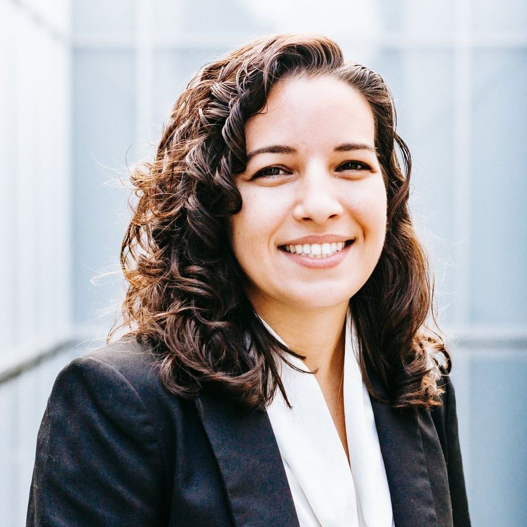 A woman with curly hair is wearing a black jacket and smiling.