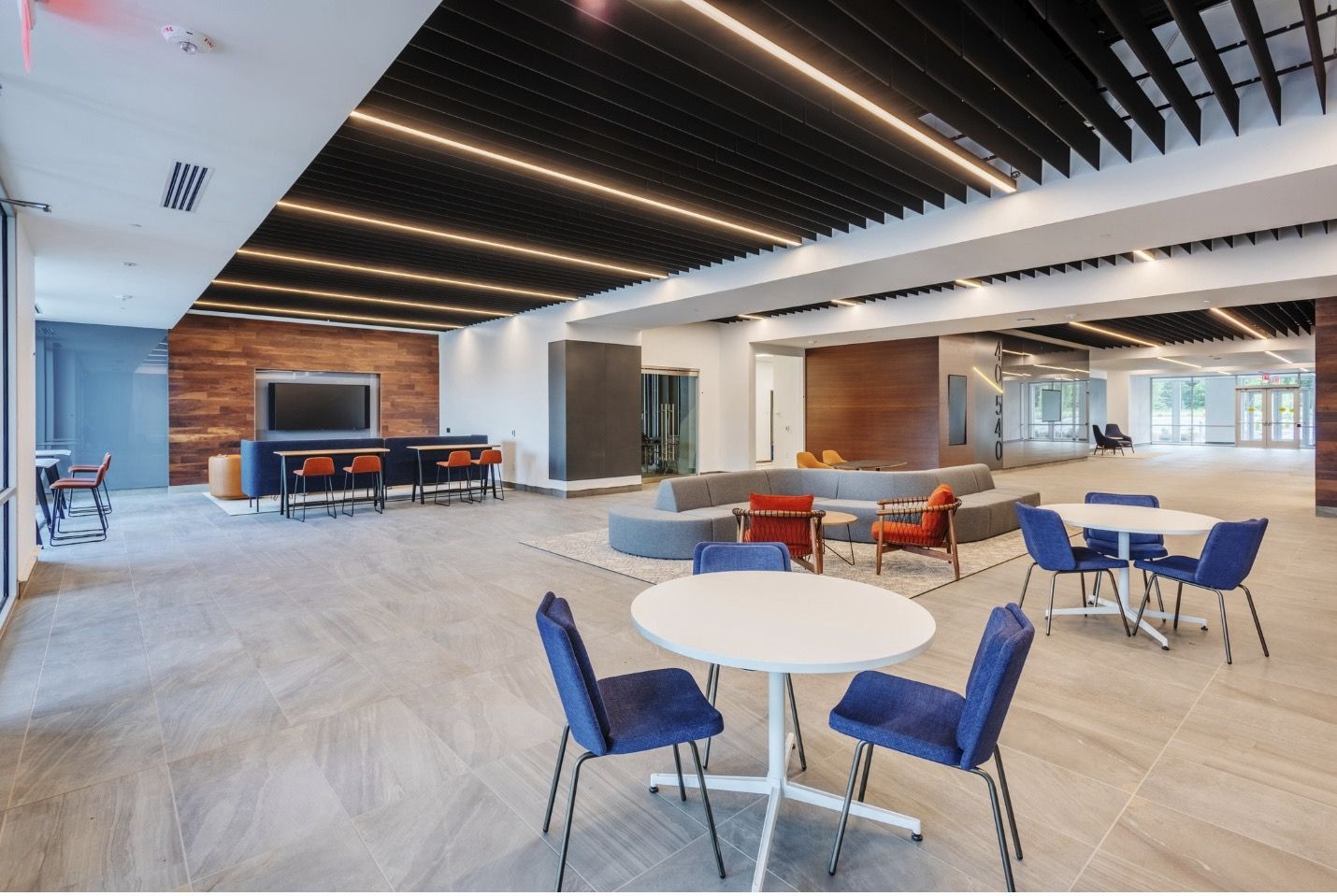 A modern, open-plan office lounge with circular white tables, blue chairs, a sectional sofa, and a wood-paneled feature wall.