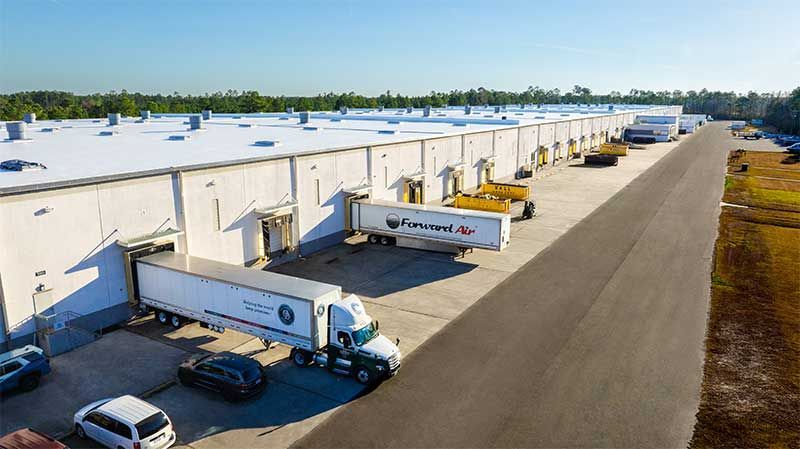 An aerial view of a large white warehouse building with several semi-truck trailers parked at the loading docks.