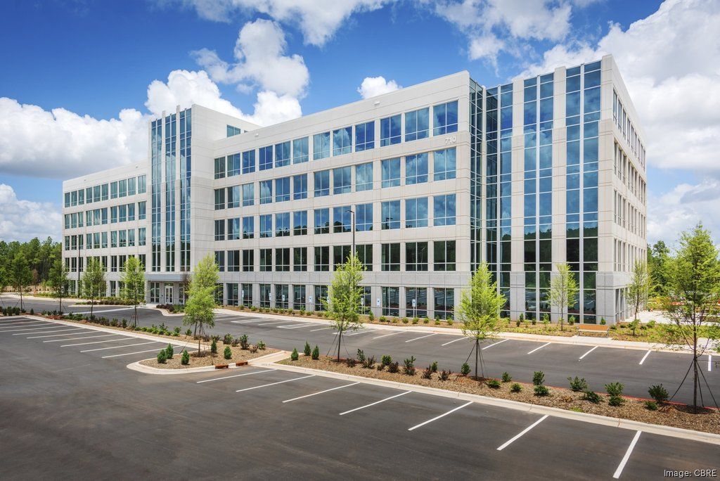 Modern multi-story office building with many windows and a large parking lot. Blue sky with clouds.
