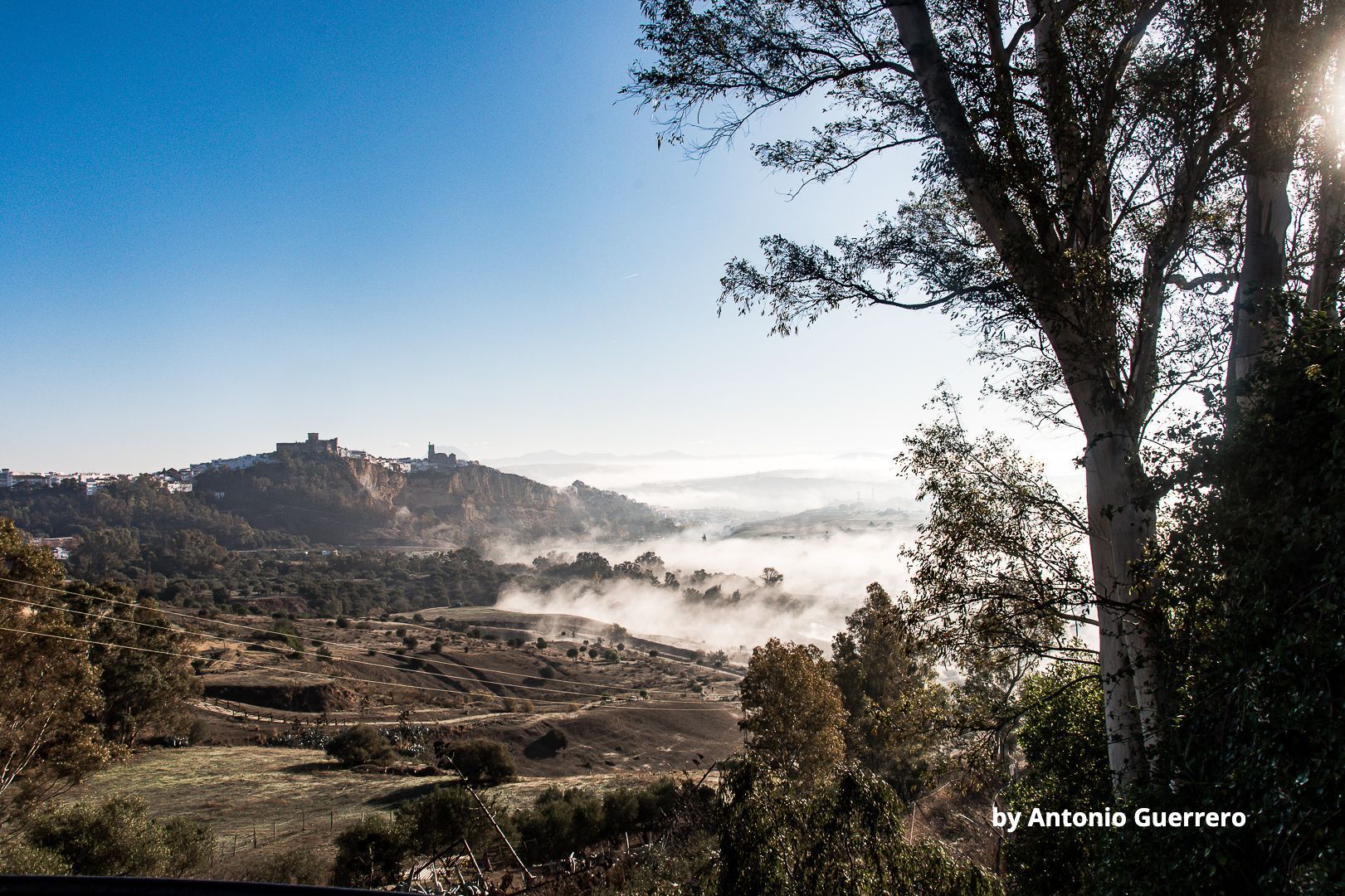 Paisaje montañoso con niebla, un pueblo distante y árboles en primer plano bajo un cielo azul brillante.