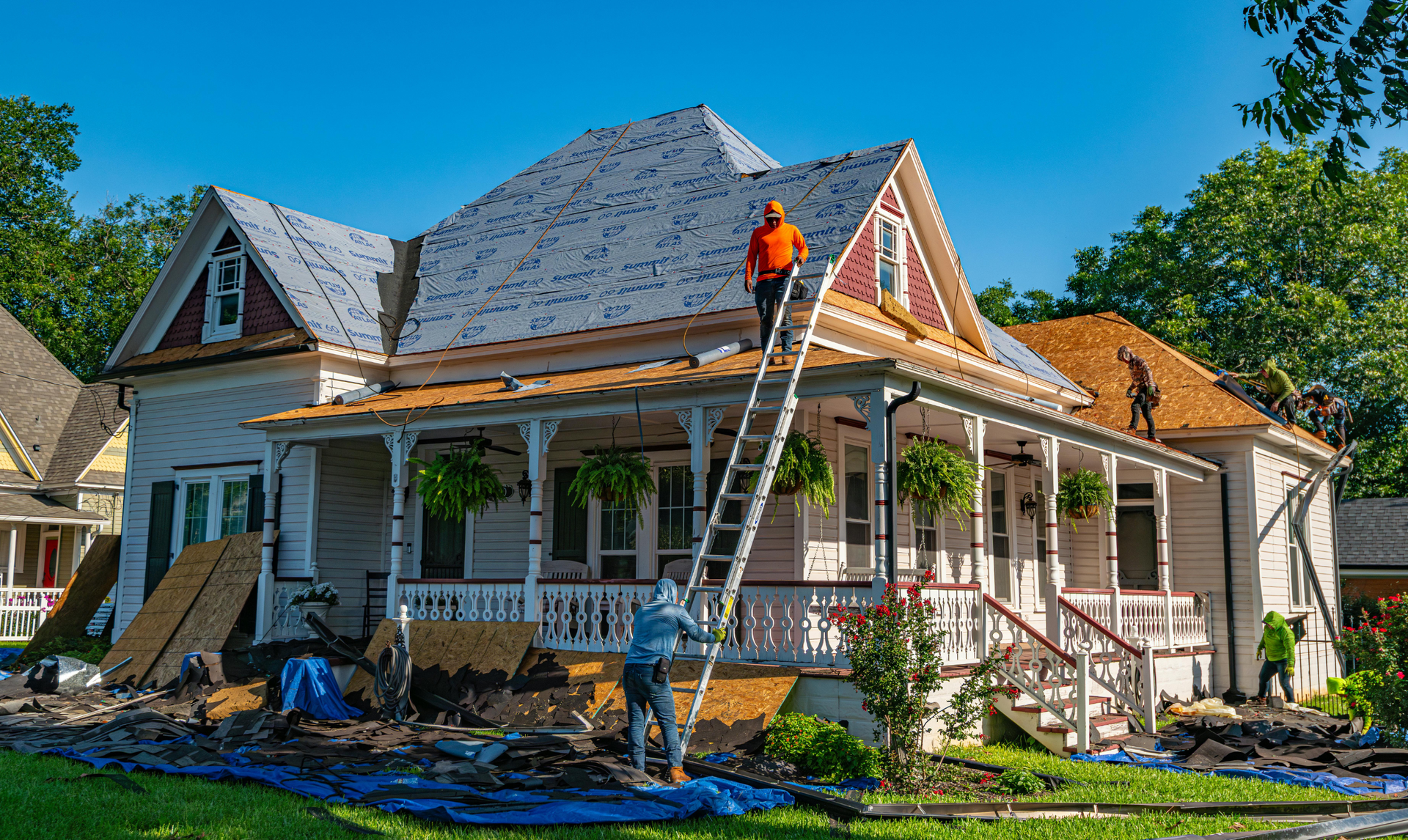 Workers replace the roof of a two-story house with a porch, using a ladder and wearing safety gear. Workers replace the roof of a two-story house with a porch, using a ladder and wearing safety gear.