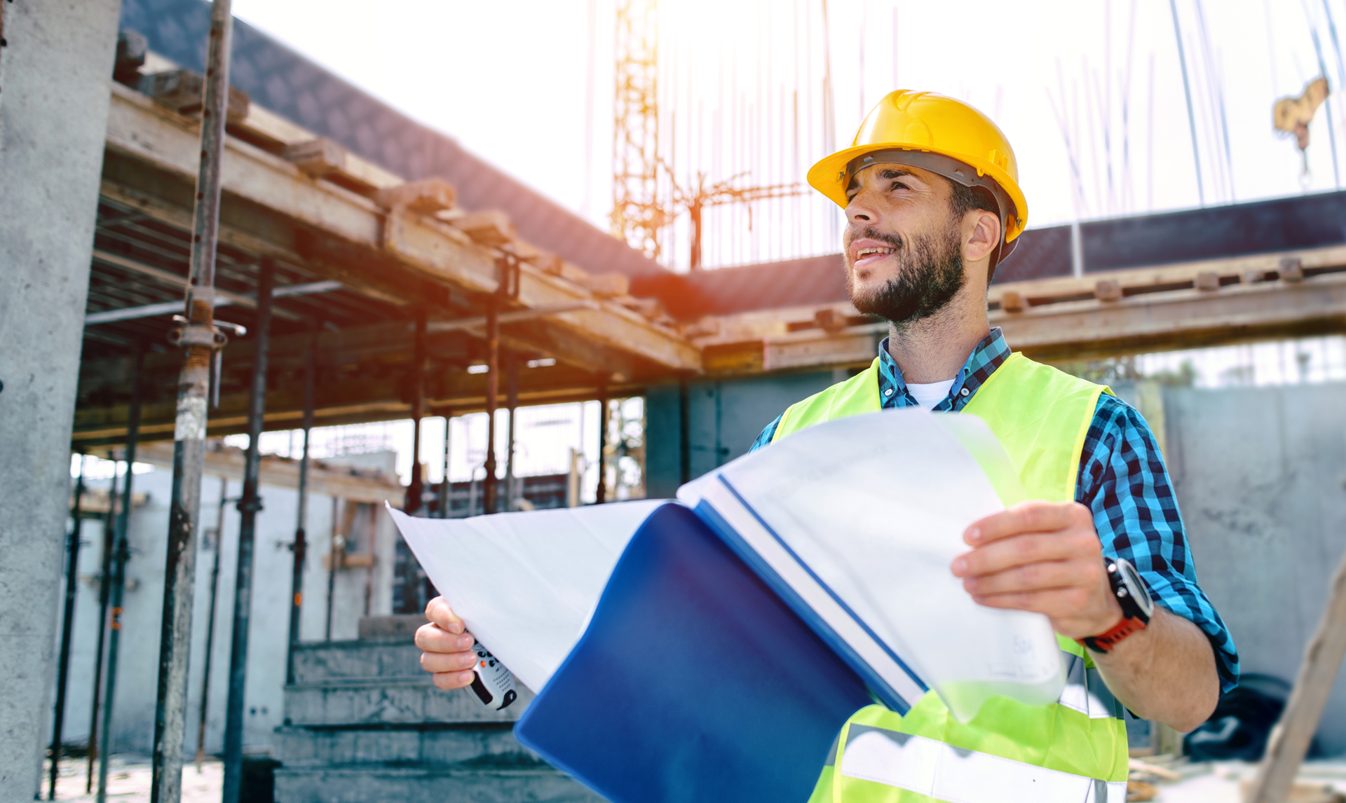 Construction worker in yellow hard hat and vest reviews blueprints at a building site.