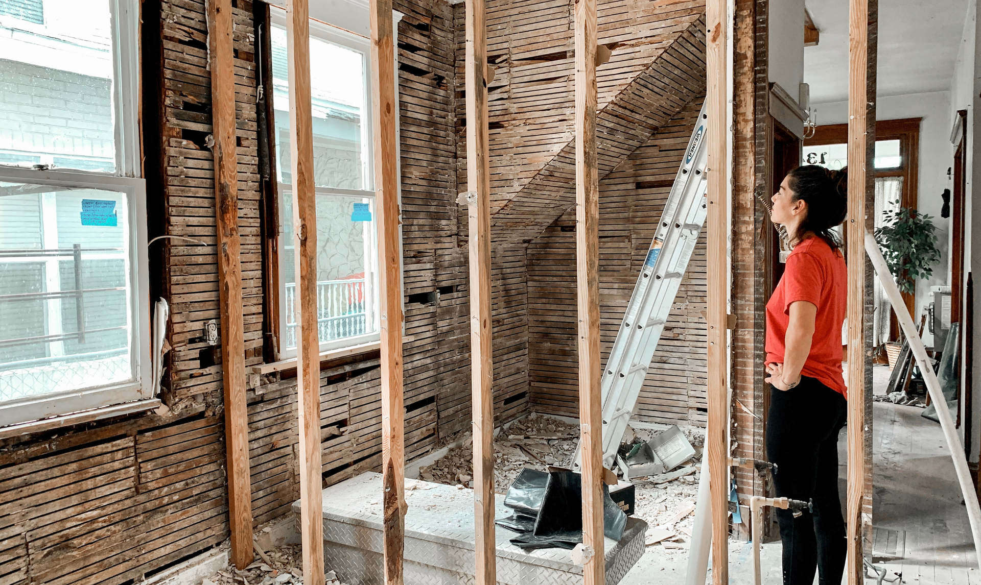 Woman in red shirt looks at a room undergoing renovation. Exposed wooden studs and plaster. Window in background.