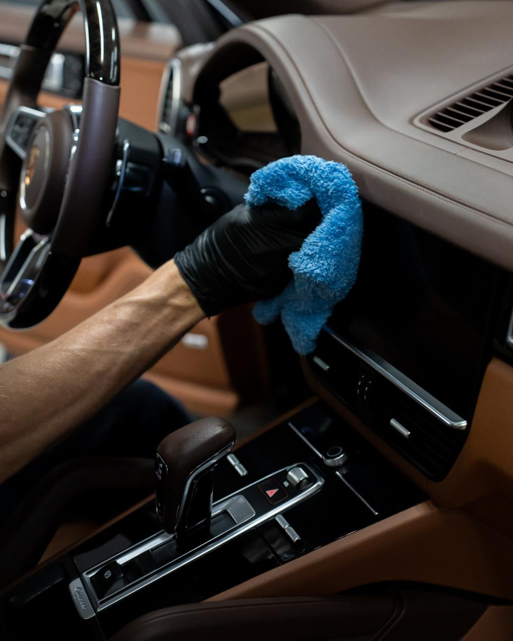 Person cleaning a car's brown leather interior with a blue microfiber cloth, wearing a black glove.