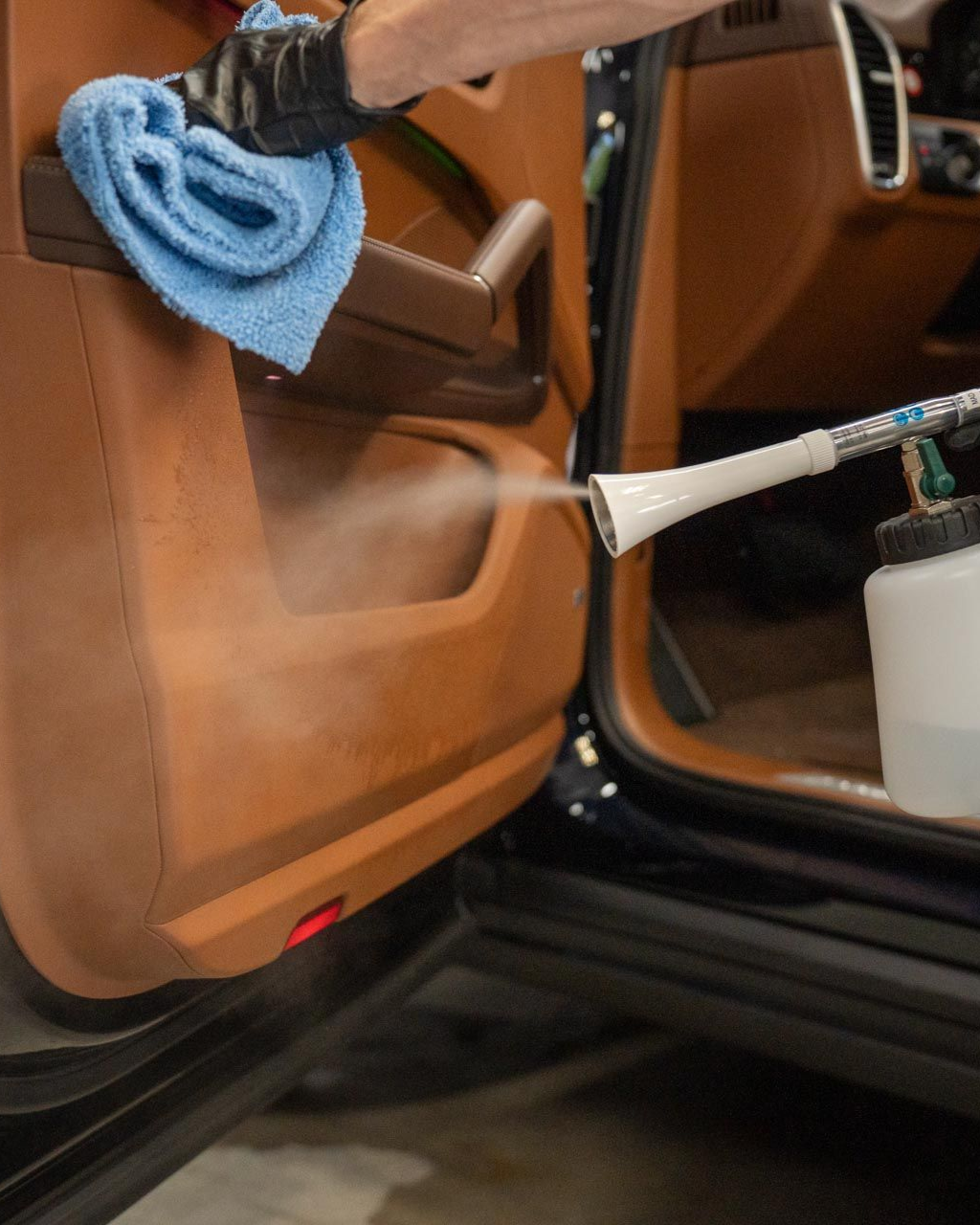 Cleaning a brown car door panel with a spray nozzle, using a blue cloth, by a gloved hand.