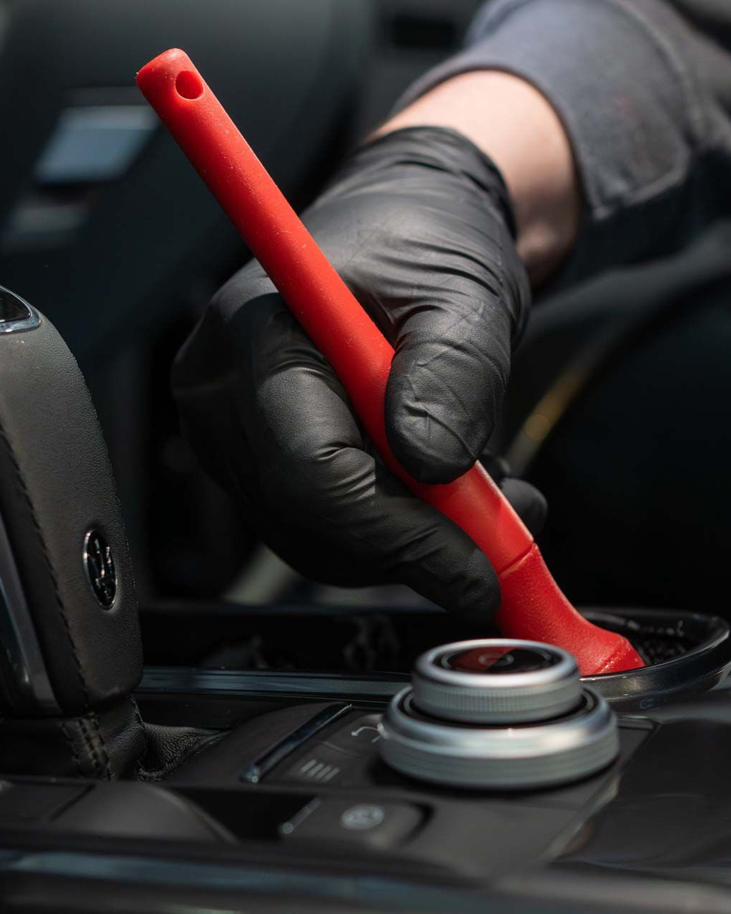 Gloved hand uses a red brush to clean a car interior, near a button and gear shift.