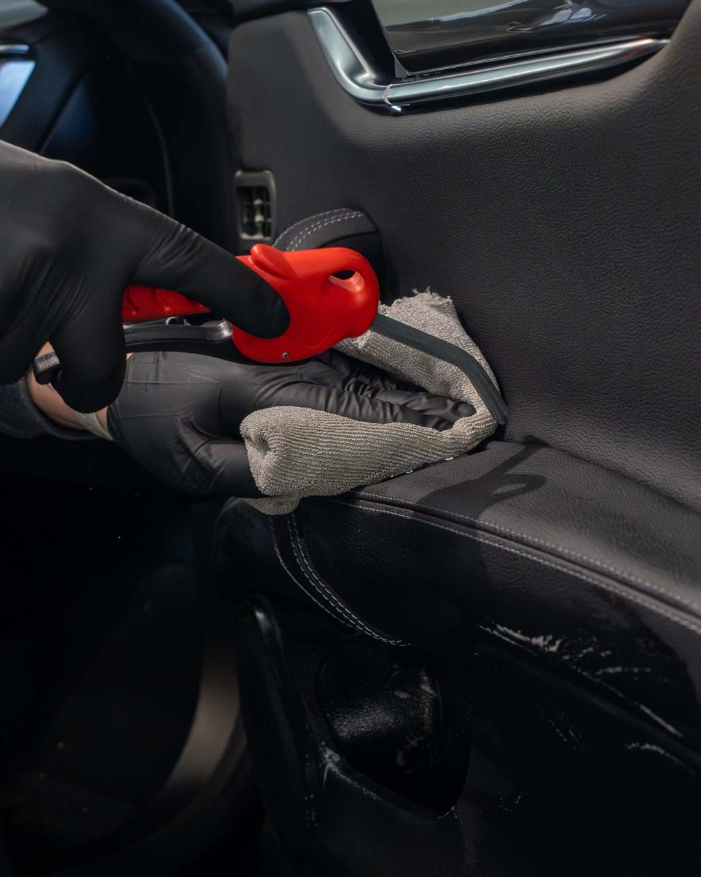 Black-gloved hands cleaning a car door panel with a red applicator and gray cloth.