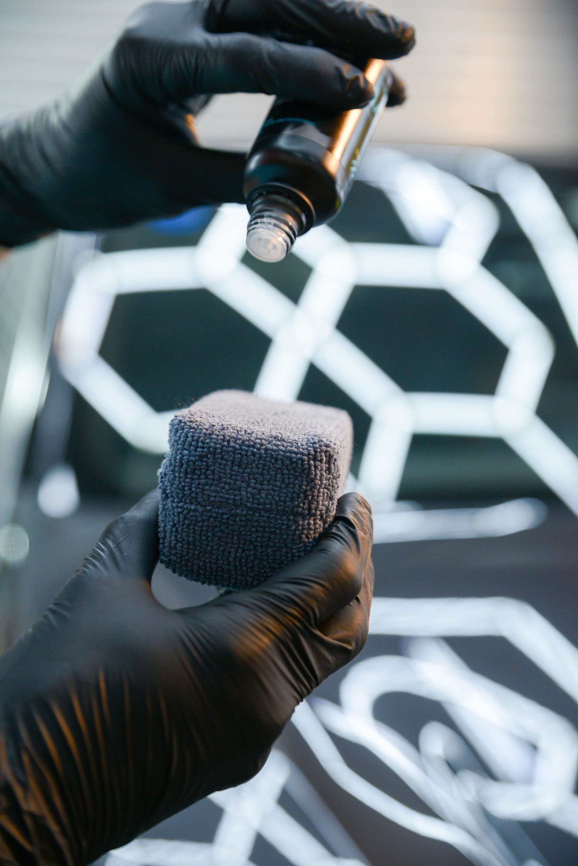 Gloved hands applying liquid from a bottle onto a blue applicator sponge for car detailing.