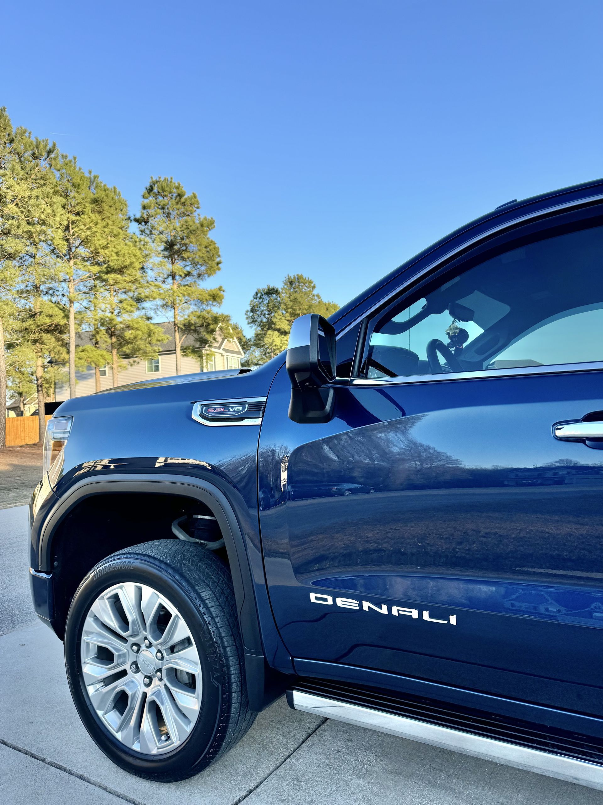 Blue GMC Denali truck parked on a concrete surface with trees and a clear blue sky in the background.