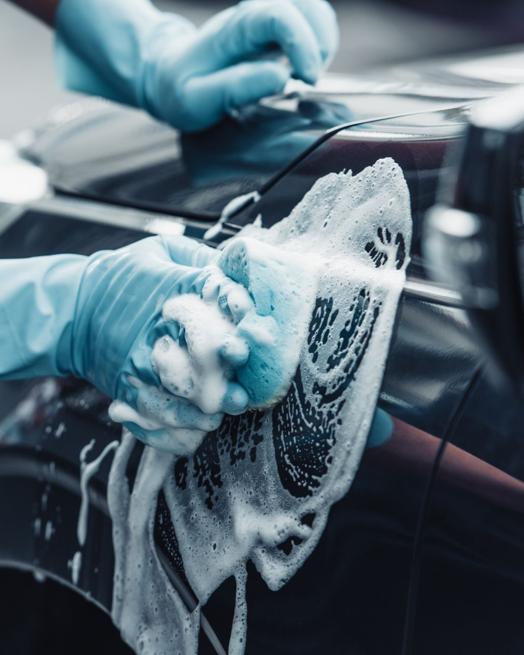 Hands in blue gloves washing a dark car with soapy sponge.