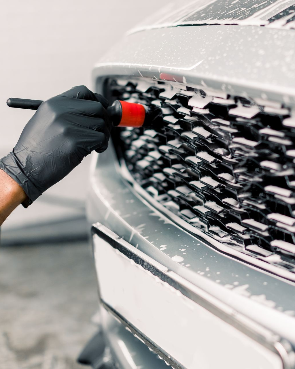 Gloved hand brushes soapy car grill, cleaning a silver car at a car wash.