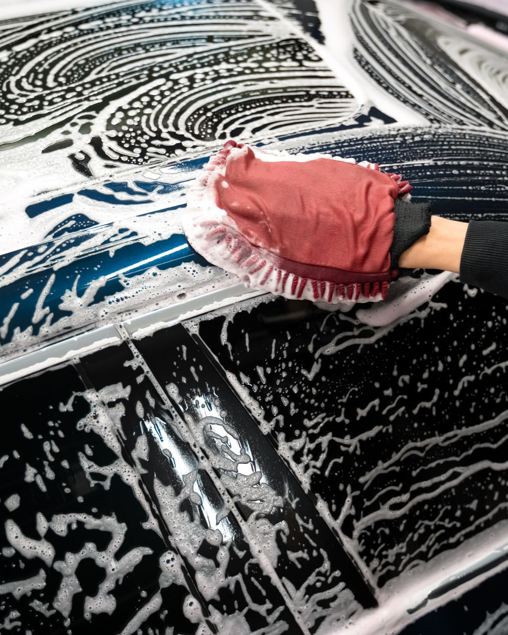 Person wearing a glove washing a blue car with a soapy mitt.