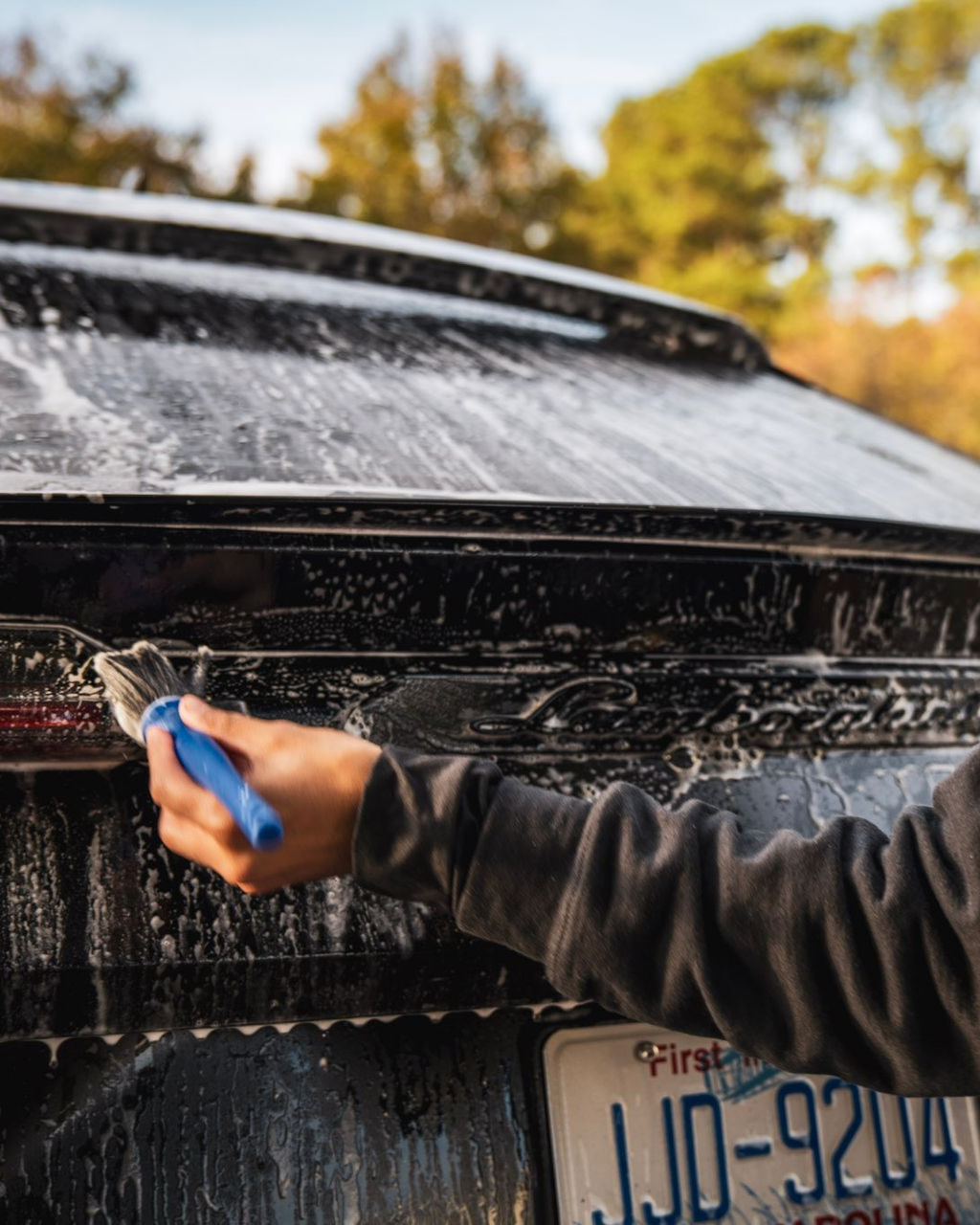 Washing a dark car with a brush. Soapy foam covers the rear; license plate visible.