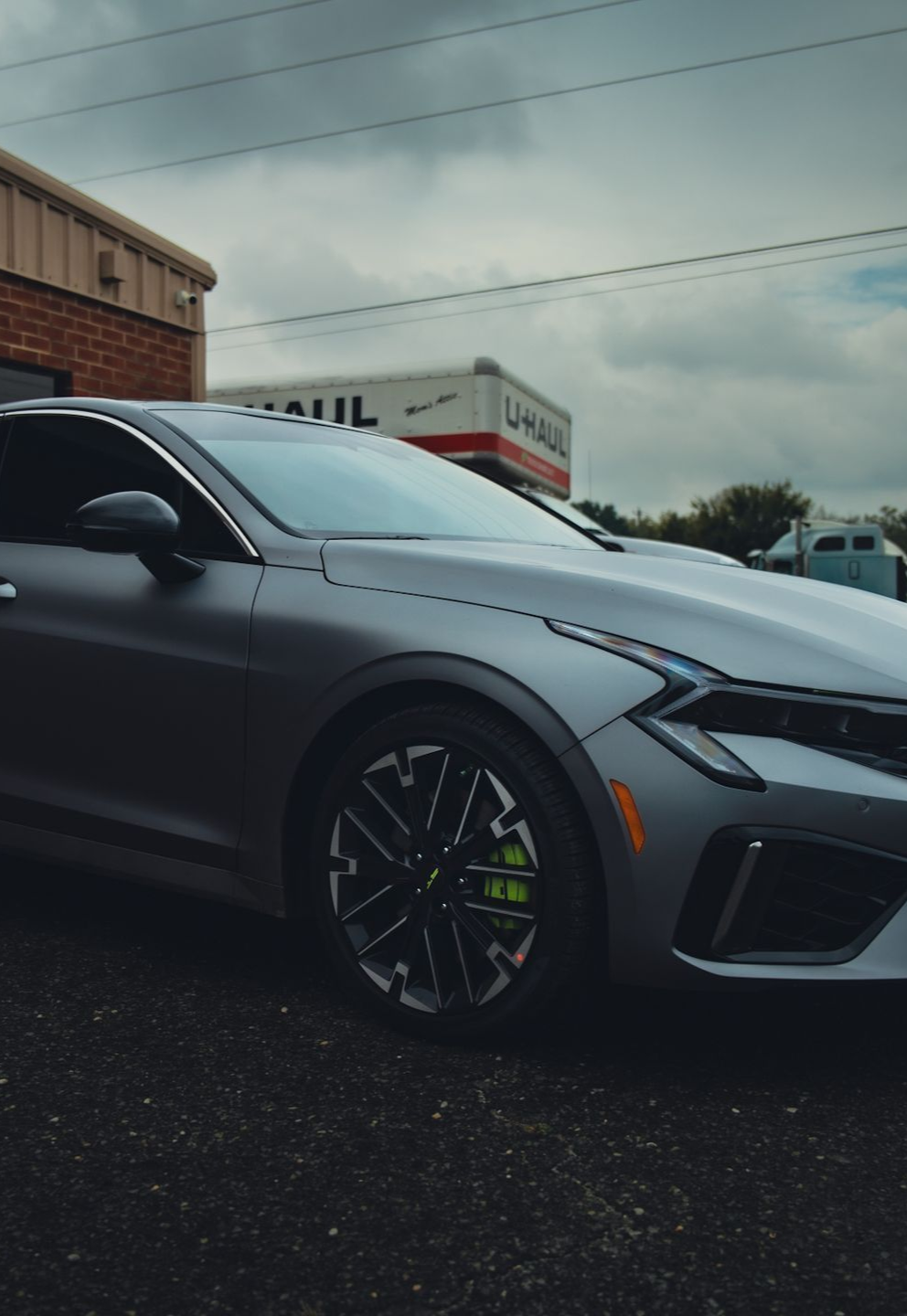 Silver Kia K5 parked near a building and U-Haul truck. Dark wheels with lime green calipers visible. Cloudy sky.