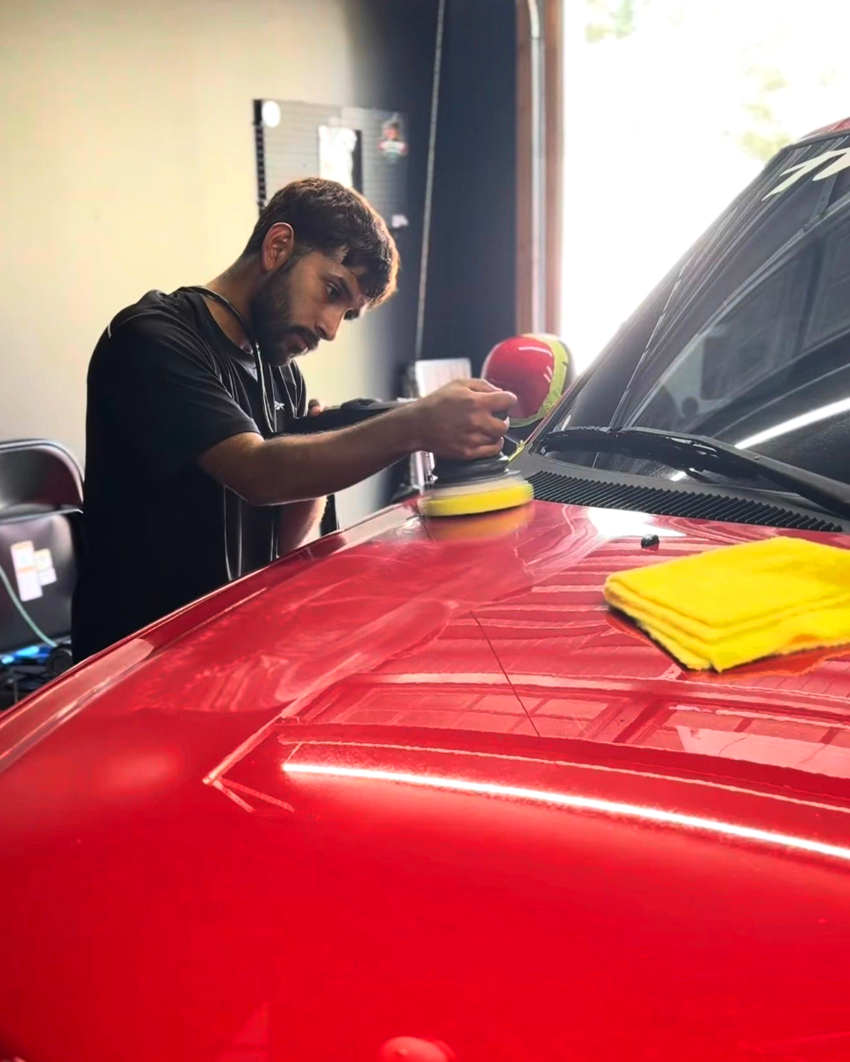 Man polishing a red car hood with an orbital buffer in a garage.