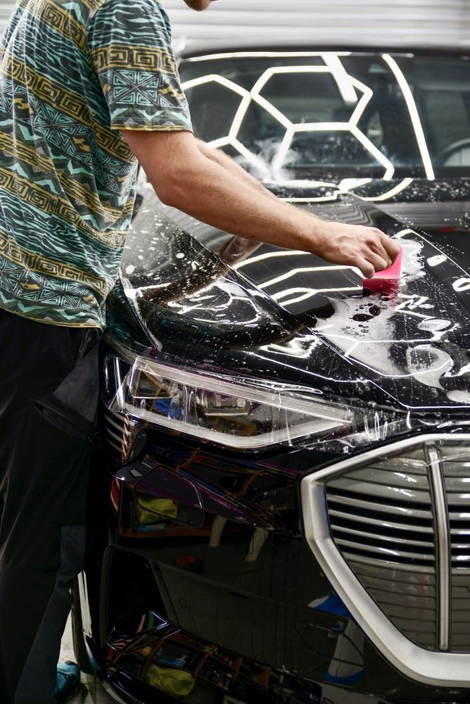 Person washing a black car with a pink sponge, bubbles visible. Shiny car hood, honeycomb lighting above.
