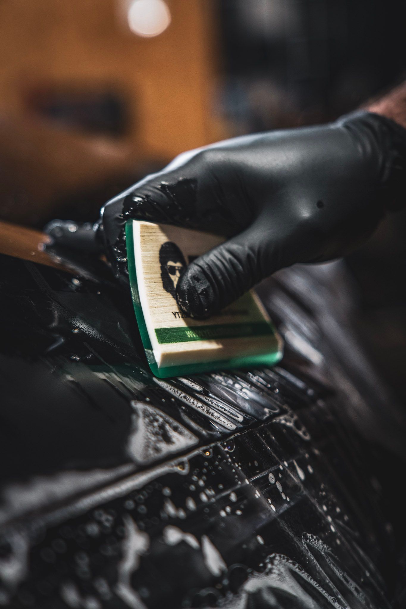 Gloved hand using a sponge on a wet, black surface, likely cleaning a car.