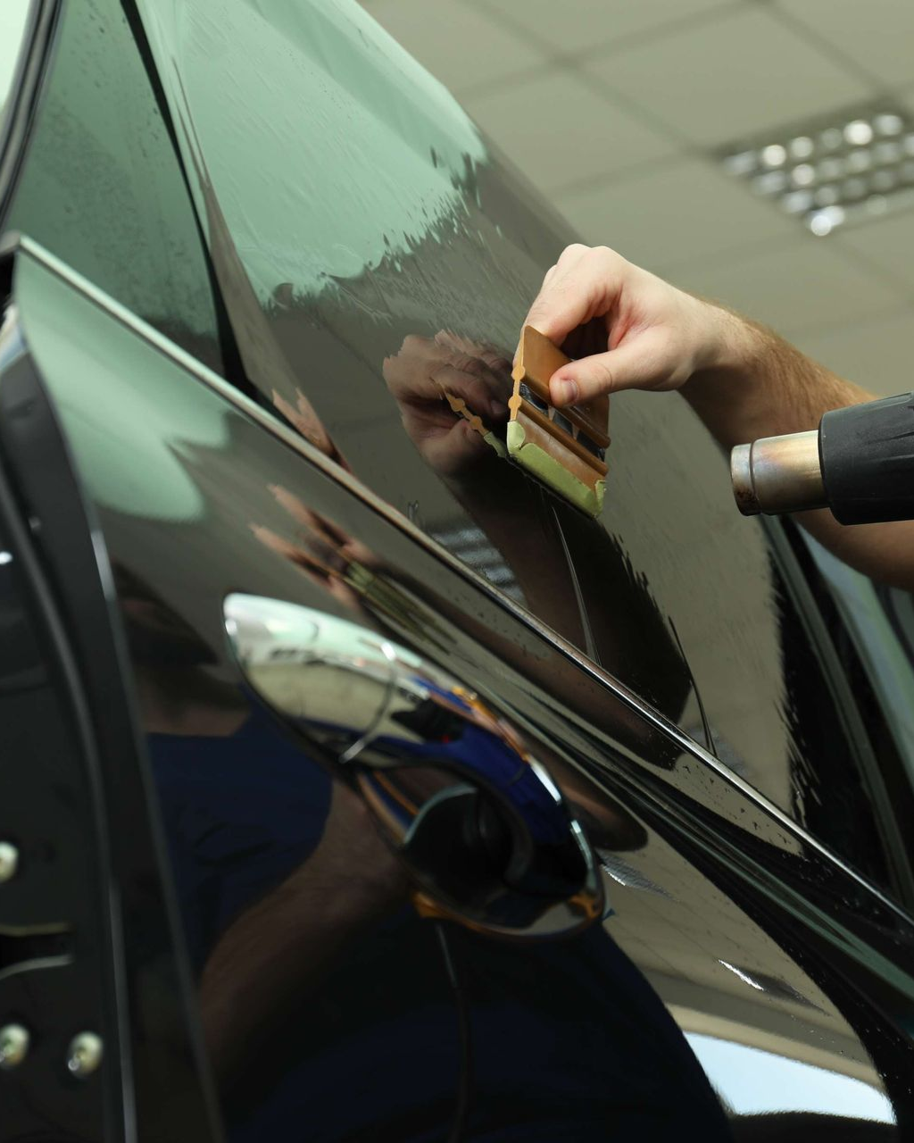 Person applying tint film to a black car door with a squeegee and heat gun.
