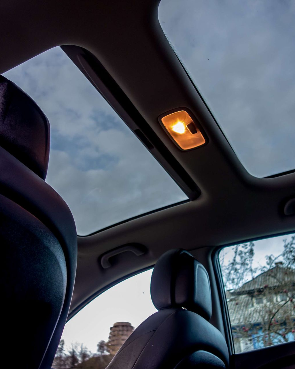 Interior car view with open sunroof, lit overhead light, and overcast sky visible.