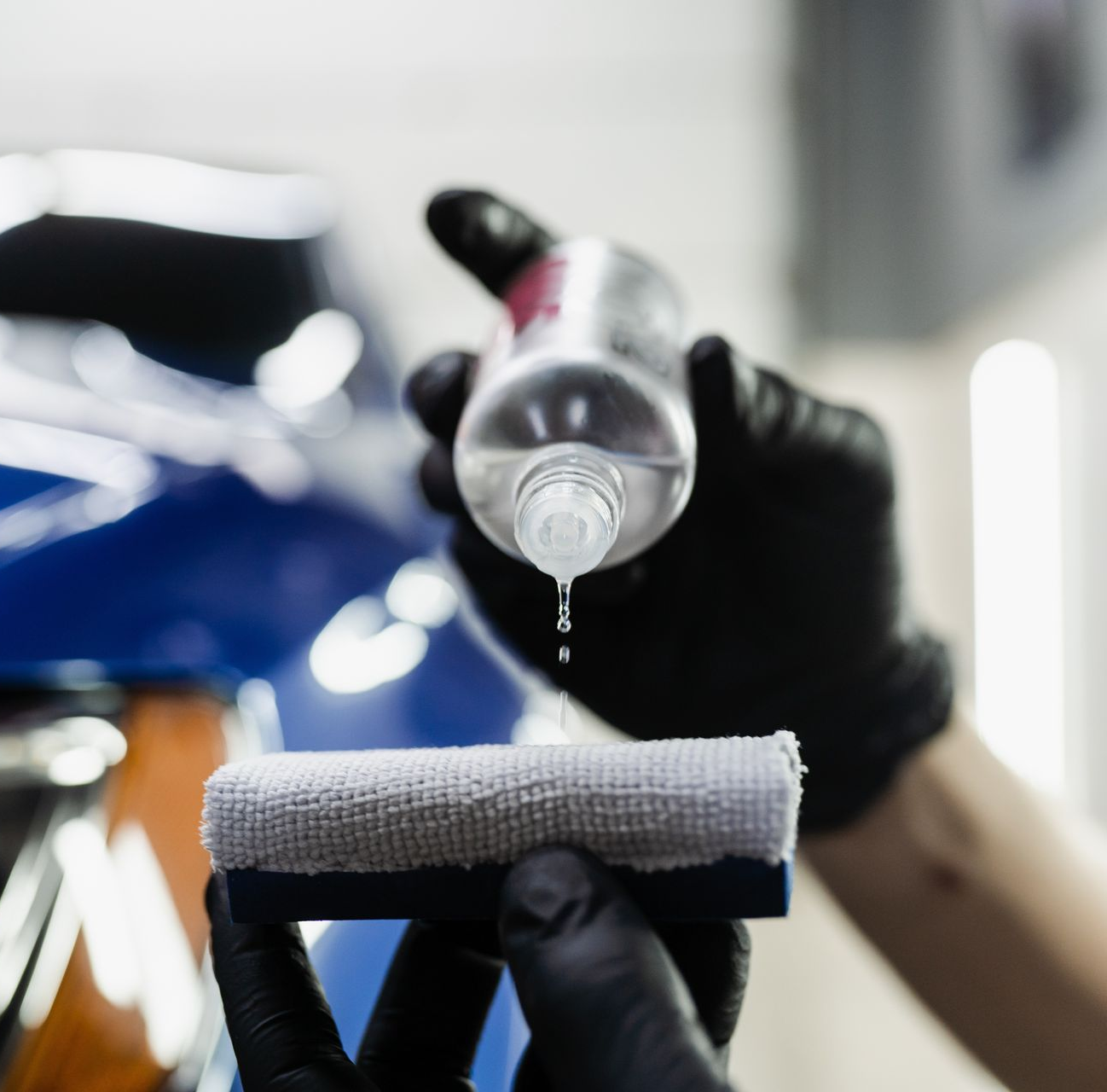 A person wearing black gloves is applying a spray to a shiny, red car in a garage.