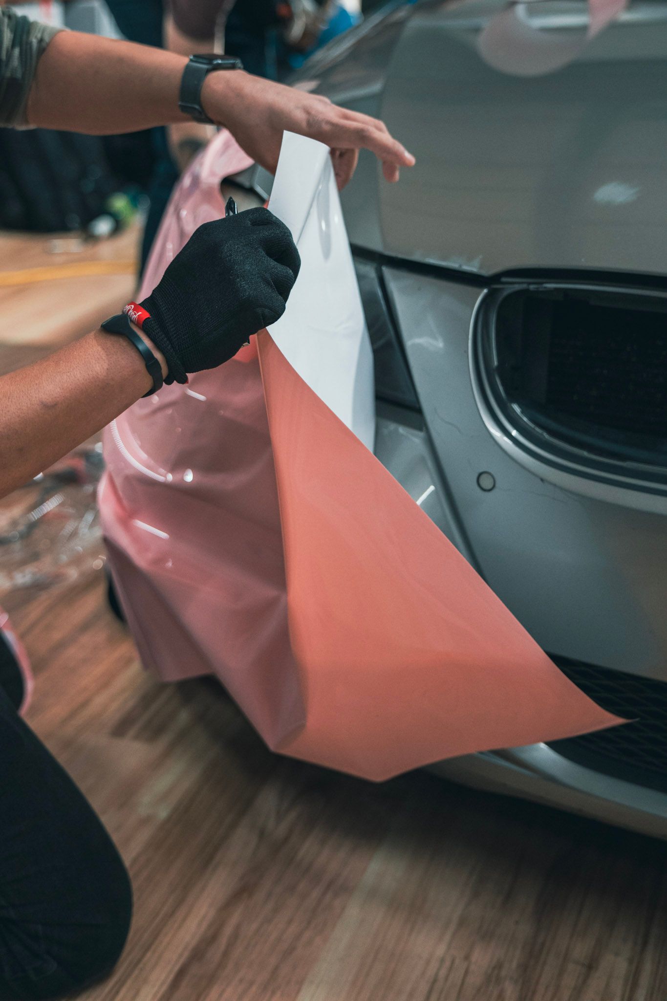 Person in black gloves peeling off pink vinyl wrap on a car bumper.