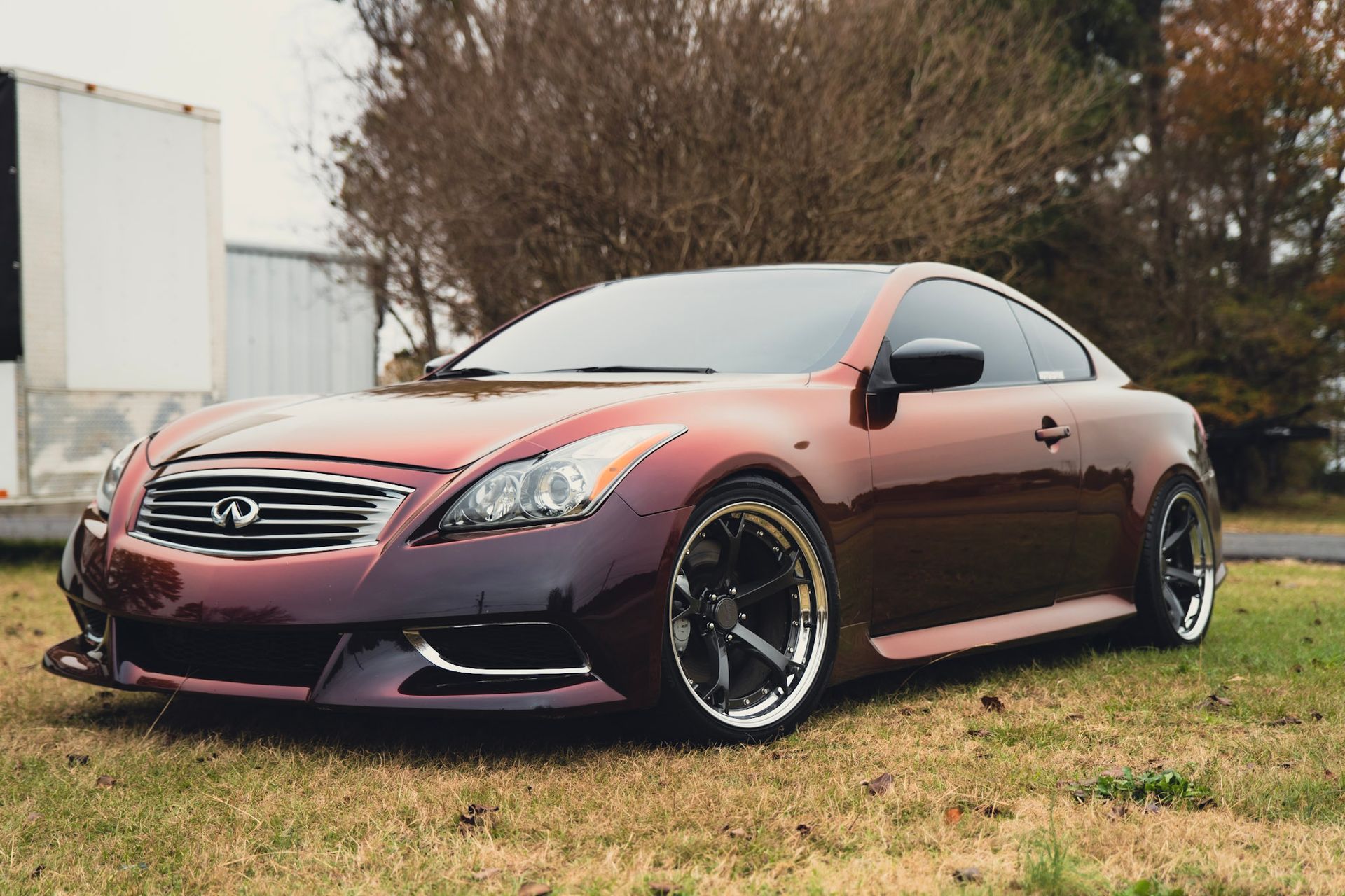 Burgundy Infiniti G37 coupe parked on grass. Black wheels, tinted windows, and a front splitter.