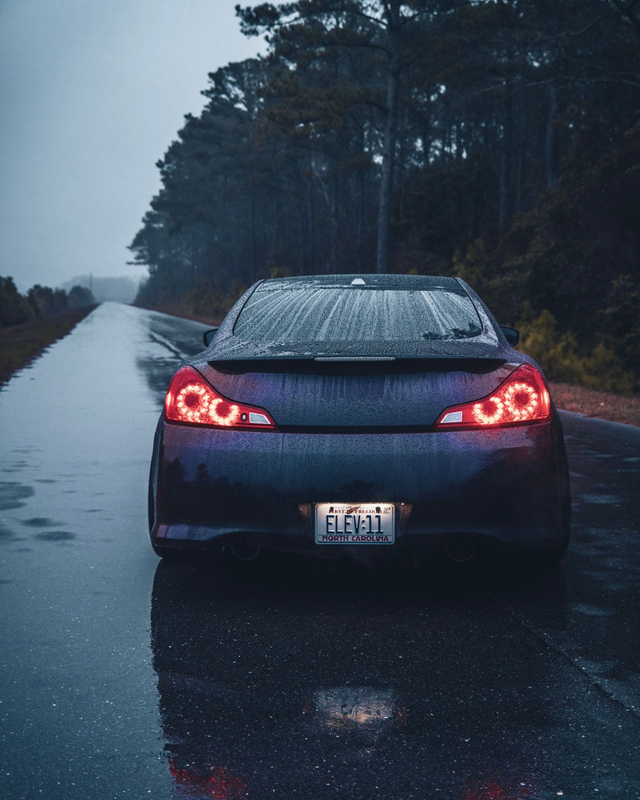 Dark car driving on a wet road in the rain, taillights glowing, surrounded by trees.