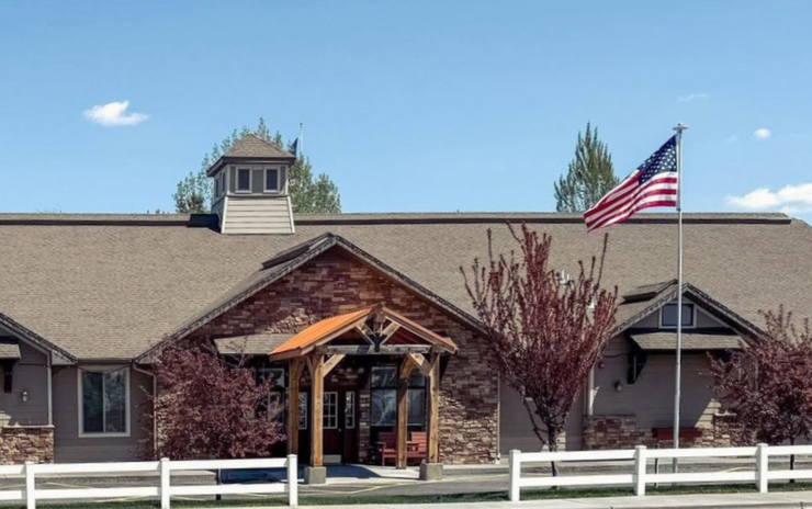 Building with flag; brown roof, stone and wood entrance, white fence, blue sky.