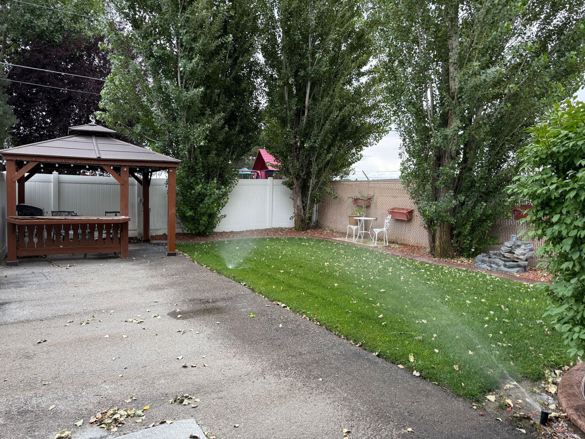 A backyard with a gazebo, lawn, and sprinkler system; trees and a fence are in the background.